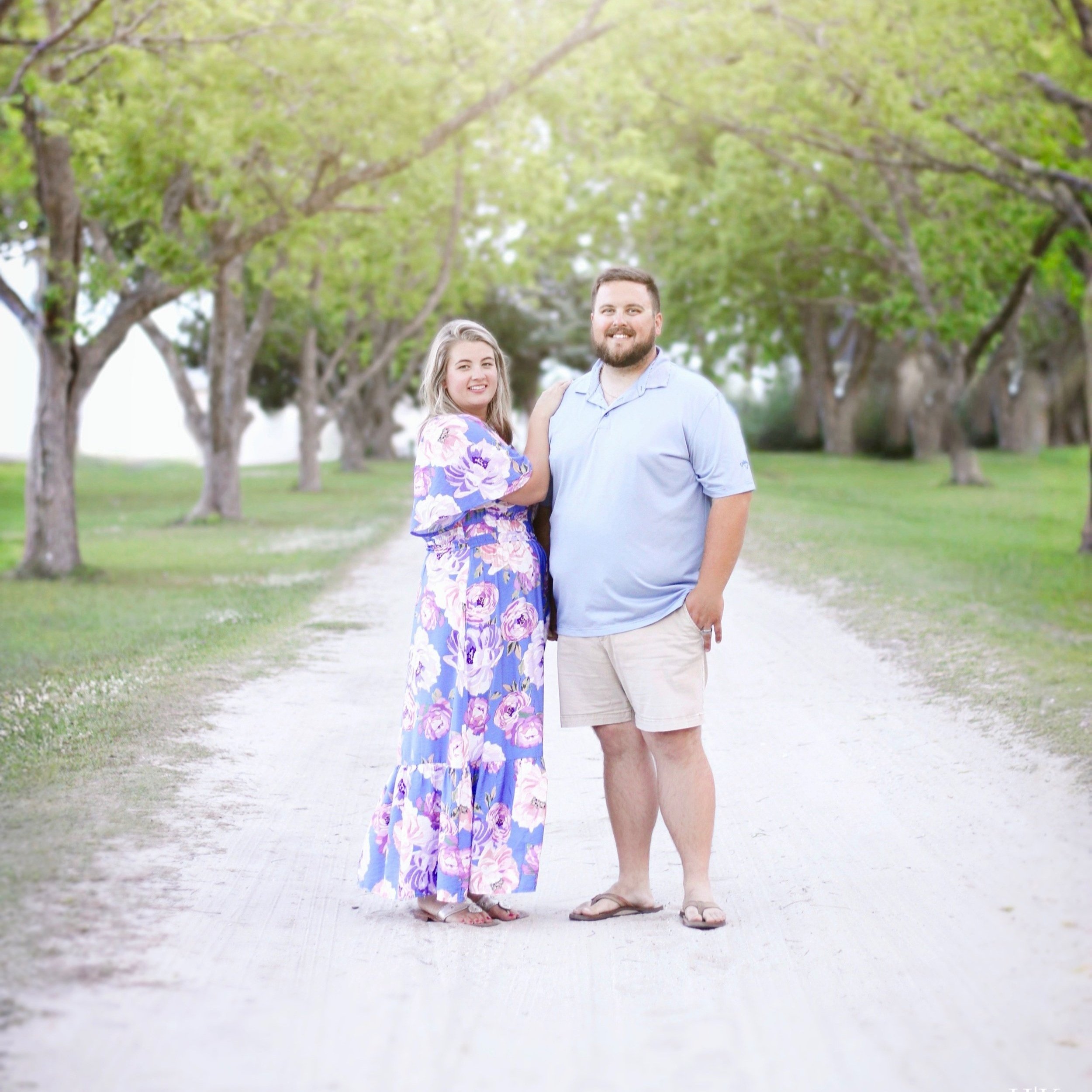 Couple standing on a tree-lined dirt path, smiling at the camera. The woman wears a floral maxi dress, while the man is dressed in a light blue polo and shorts. The background features lush green trees and a serene atmosphere, ideal for outdoor photography.