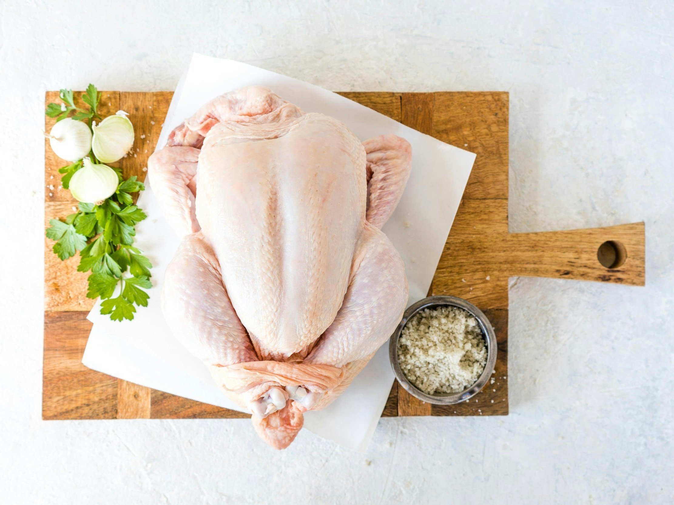 Raw whole chicken on a wooden cutting board, accompanied by fresh parsley, garlic cloves, and a bowl of salt, ideal for meal preparation or cooking.